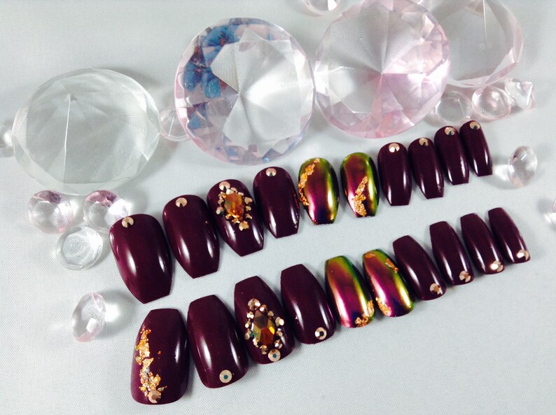 Puede incluir: U&ntilde;as postizas de ata&uacute;d color burdeos con detalles dorados y pedrer&iacute;a. Algunas u&ntilde;as tienen un acabado hologr&aacute;fico.