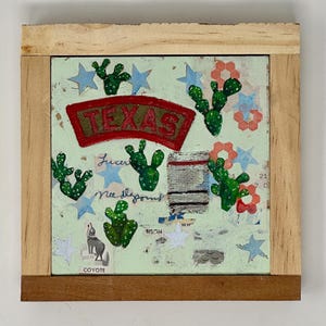 Peut inclure: Œuvre d'art mixte avec un fond vert clair, un emblème Texas rouge et vert, des cactus verts, des étoiles bleues et une illustration de coyote. L'œuvre est encadrée en bois naturel.