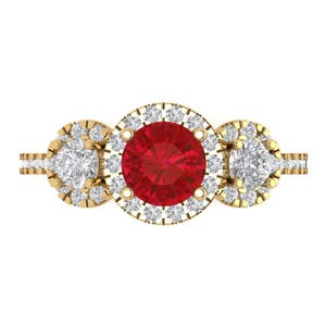 May include: A gold ring featuring a large, round, vibrant red centre stone surrounded by a halo of small, clear, sparkling stones. Two smaller, round, clear stones are set on either side, each also encircled by a halo of tiny clear stones.