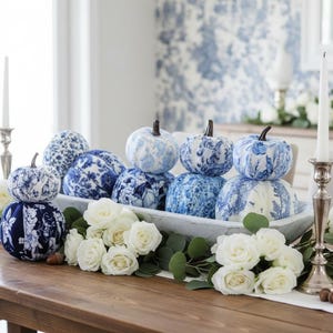 Könnte beinhalten: Eine Sammlung von Stoffkürbissen mit verschiedenen blau-weißen Blumenmustern, die in einer langen, weißen, rechteckigen Holzschale angeordnet sind. Das Arrangement wird durch weiße Rosen, grüne Eukalyptusblätter und silberne Kerzenständer ergänzt.