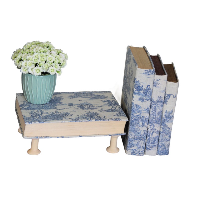 Puede incluir: Un soporte con forma de libro con estampado toile azul y blanco y cuatro peque&ntilde;as patas de madera, que sostiene una maceta de cer&aacute;mica azul claro acanalada con flores blancas, junto a dos libros verticales a juego con la misma tela.