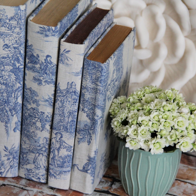 Puede incluir: Tres libros con cubiertas de color beige claro con un patr&oacute;n azul de toile de jouy de figuras y paisajes, colocados junto a una maceta de cer&aacute;mica verde claro llena de peque&ntilde;as flores de color verde p&aacute;lido con muchos p&eacute;talos.