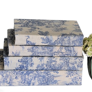 Puede incluir: Una pila de cuatro libros o cajas rectangulares cubiertos con una tela de color beige claro con un patr&oacute;n azul de toile de jouy. El estampado muestra escenas pastorales detalladas con figuras, &aacute;rboles y aves. Los objetos est&aacute;n apilados.
