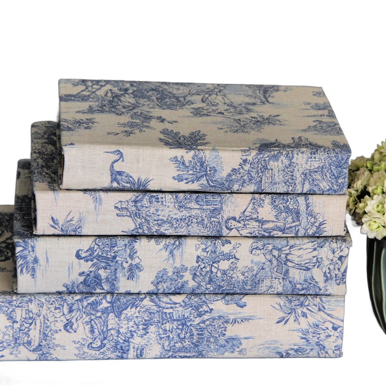Puede incluir: Una pila de cuatro libros o cajas rectangulares cubiertos con una tela de color beige claro con un patr&oacute;n azul de toile de jouy. El estampado muestra escenas pastorales detalladas con figuras, &aacute;rboles y aves. Los objetos est&aacute;n apilados.