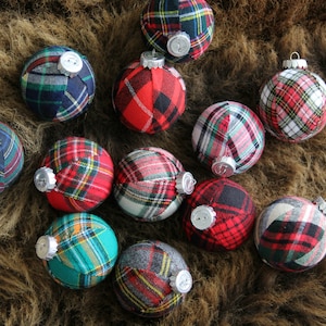 May include: A collection of 12 Christmas ornaments made from plaid fabric in various colors, including red, green, blue, and white. The ornaments are arranged on a brown furry surface.