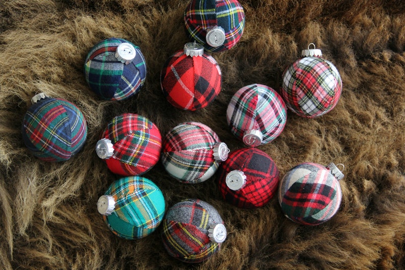 May include: A collection of 12 Christmas ornaments made from plaid fabric in various colors, including red, green, blue, and white. The ornaments are arranged on a brown furry surface.