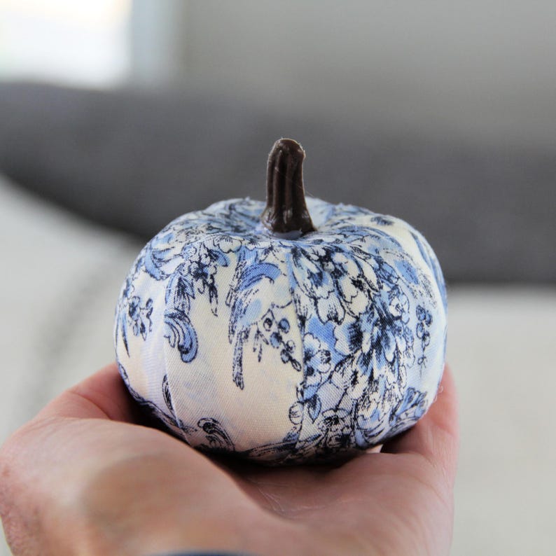 Pode incluir: Uma pequena ab&oacute;bora decorativa coberta de tecido com uma base branca e um padr&atilde;o floral azul estilo toile. A ab&oacute;bora tem um caule texturizado castanho-escuro no topo. O objeto &eacute; esf&eacute;rico com segmentos verticais e aspeto almofadado.