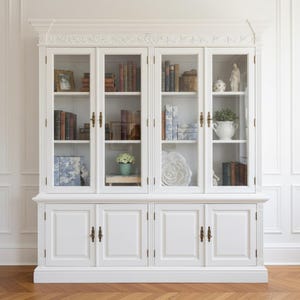 Puede incluir: Una gran alacena de madera blanca con cuatro puertas superiores de cristal y herrajes de lat&oacute;n, que muestra estantes con libros, figuras decorativas y objetos con motivos azules y blancos. La base incluye cuatro puertas blancas s&oacute;lidas.