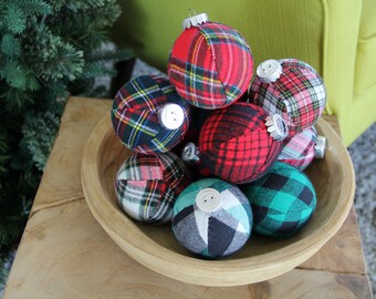 May include: A wooden bowl filled with plaid Christmas ornaments. The ornaments are round and covered in red, green, and blue plaid fabric. Each ornament has a silver cap and a white button. The bowl sits on a wooden table.