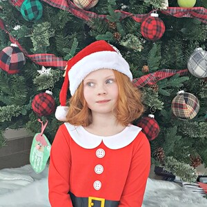 May include: A young girl wearing a red Santa Claus costume with a white collar and black and white plaid pants sits in front of a Christmas tree decorated with plaid ornaments.