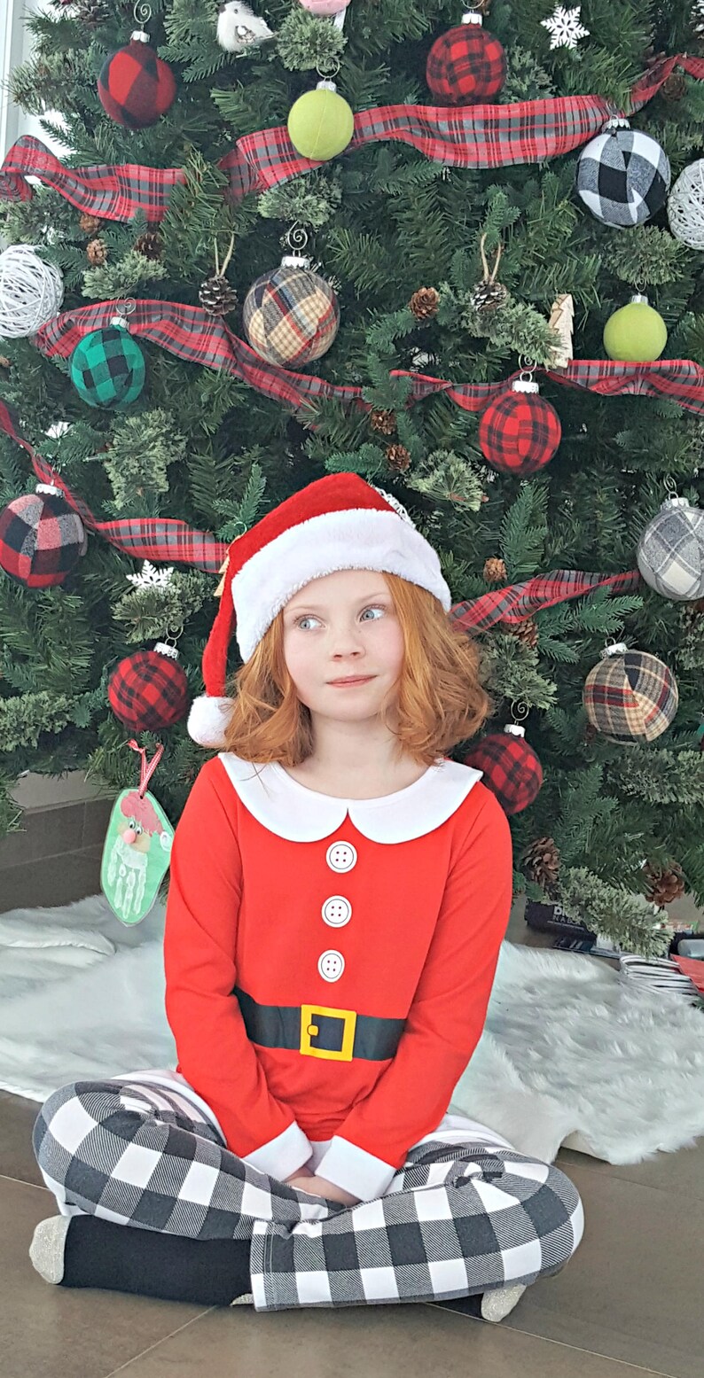May include: A young girl wearing a red Santa Claus costume with a white collar and black and white plaid pants sits in front of a Christmas tree decorated with plaid ornaments.