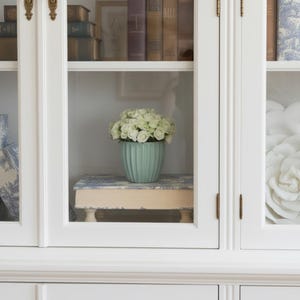 Puede incluir: Una maceta de cer&aacute;mica de color verde claro con estr&iacute;as verticales llena de rosas blancas, apoyada sobre un soporte de madera rectangular con un patr&oacute;n floral azul y blanco, todo expuesto en un armario blanco con puertas de cristal y herrajes de lat&oacute;n.