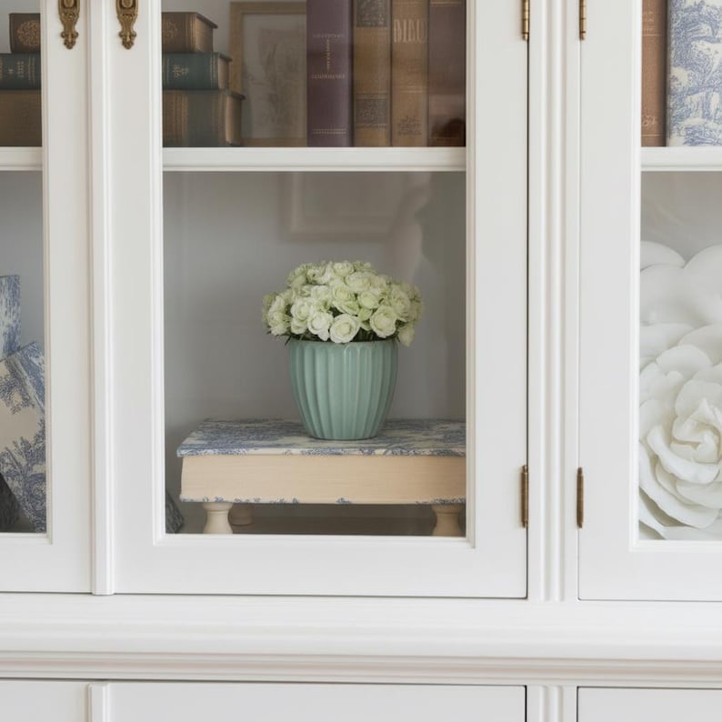Puede incluir: Una maceta de cer&aacute;mica de color verde claro con estr&iacute;as verticales llena de rosas blancas, apoyada sobre un soporte de madera rectangular con un patr&oacute;n floral azul y blanco, todo expuesto en un armario blanco con puertas de cristal y herrajes de lat&oacute;n.
