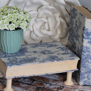Puede incluir: Un soporte rectangular con forma de libro cubierto de tela toile azul y blanca con escenas pastorales, apoyado sobre cuatro patas de madera clara en forma de carrete. Una maceta de cer&aacute;mica verde claro acanalada contiene flores blancas.