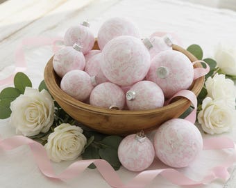 Puede incluir: Una colección de adornos navideños redondos con un patrón floral toile en rosa suave y blanco. Los adornos se exhiben en un cuenco de madera, acentuados por rosas blancas, hojas de eucalipto verdes y una cinta de color rosa claro.