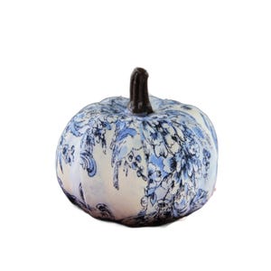 Pode incluir: Um ornamento de ab&oacute;bora decorativo com um exterior de tecido branco e um padr&atilde;o floral azul tipo toile. A ab&oacute;bora tem uma forma arredondada e segmentada com um caule texturizado castanho escuro que sobressai do centro do topo do objeto.