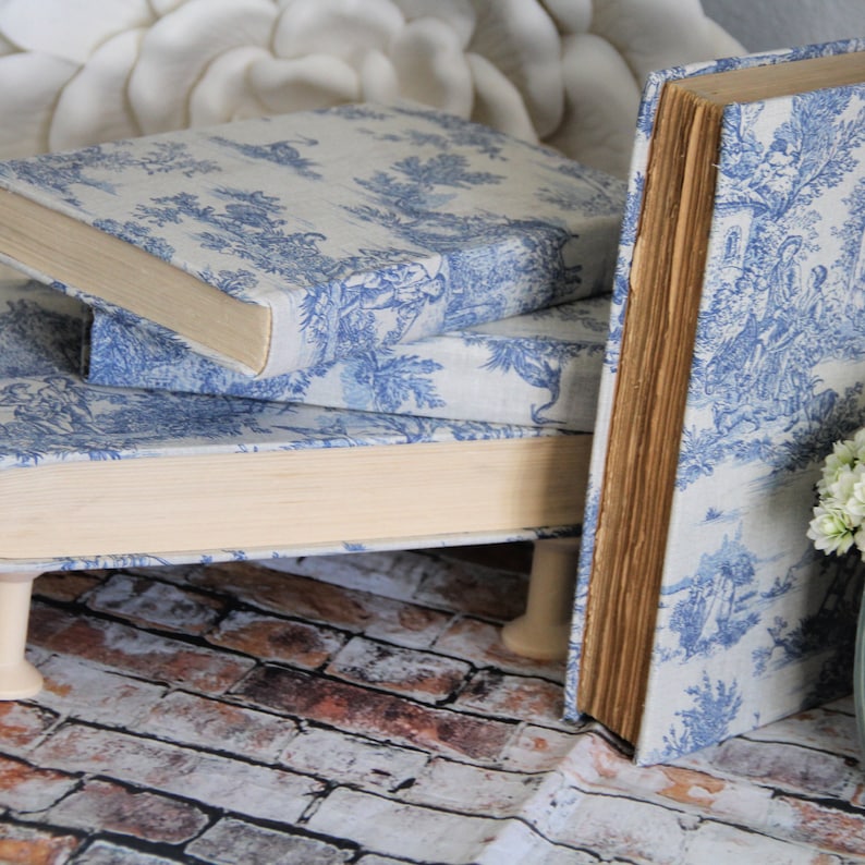 Puede incluir: Una pila de tres libros con una cubierta de tela con estampado toile en azul y blanco. Los libros est&aacute;n elevados sobre peque&ntilde;as patas de madera con forma de carrete. Las p&aacute;ginas tienen un aspecto envejecido de color tostado claro y las cubiertas muestran escenas pastorales.