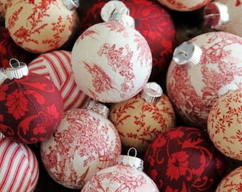 May include: A collection of round Christmas ornaments featuring various fabric patterns in red and cream. The designs include toile scenes, floral motifs, and striped prints. Each ornament is topped with a silver metal cap for hanging.