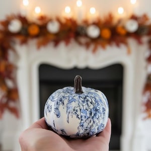 Pode incluir: Uma pequena ab&oacute;bora decorativa com base de tecido branco, um padr&atilde;o floral azul tipo toile e um caule castanho escuro. A ab&oacute;bora tem uma forma arredondada e segmentada, exibindo uma estampa bot&acirc;nica azul detalhada no tecido.