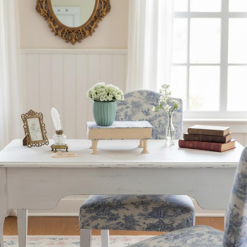Puede incluir: Un escritorio de madera blanca con un jarr&oacute;n acanalado de color verde claro con flores blancas, un peque&ntilde;o marco dorado, una pluma blanca en un tintero de cristal, un soporte con forma de libro y una pila de tres libros con cubiertas azules y marrones.
