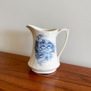 May include: A white ceramic jug with a blue and white floral design. The jug has a handle and a spout. It is sitting on a wooden table.