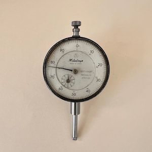 May include: A white dial analog dial gauge with a black bezel and a silver stem. The gauge reads from 0 to 100 and has the text "Mitutoyo MADE IN JAPAN" and "001-1.000" NO 2416-10 JEWELED" printed on the face.