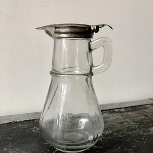 May include: A clear glass pitcher with a silver lid and a handle. The pitcher has a textured, ribbed design.