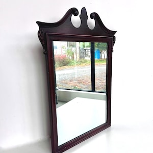 May include: A rectangular, dark cherry-stained wooden mirror with a decorative top. The mirror reflects the surrounding environment. The frame is a deep red-brown colour, and the mirror is angled against a white wall.