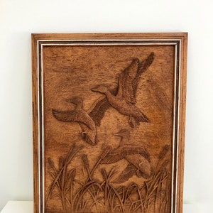 May include: A wooden wall hanging featuring a carved image of three ducks in flight over a field of tall grass. The wood is a dark brown colour and the carving is detailed.