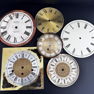 May include: A collection of vintage clock faces in various shapes and sizes. The faces include Roman numerals and the brand name "Elgin Quartz". Colors include gold, white, and brown. The clock faces are likely for restoration or crafting projects.