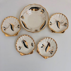 May include: Set of five white ceramic dishes with gold trim and black floral designs. The dishes are shaped like seashells.