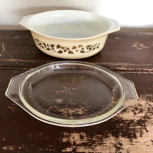 May include: A white casserole dish with a gold leaf design and a clear glass lid. The dish is on a wooden surface.