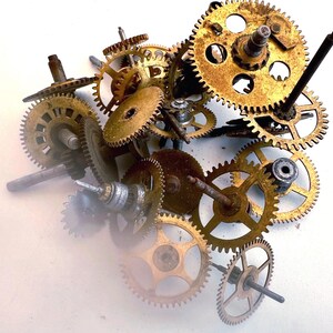 May include: A collection of vintage clock gears in various sizes and shapes. The gears are made of brass and have a worn, aged appearance. Some gears have attached rods and shafts, and the arrangement is scattered on a white surface.
