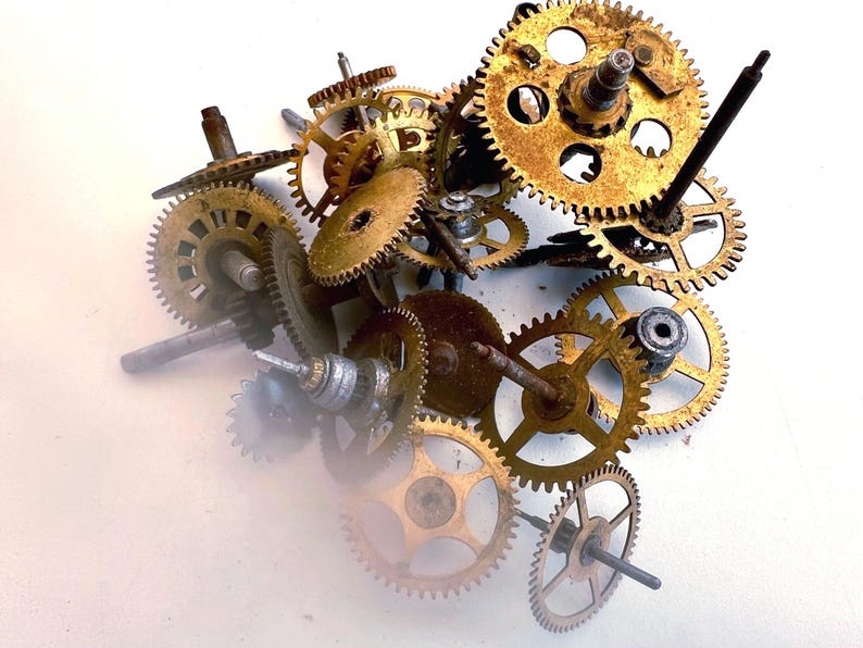 May include: A collection of vintage clock gears in various sizes and shapes. The gears are made of brass and have a worn, aged appearance. Some gears have attached rods and shafts, and the arrangement is scattered on a white surface.