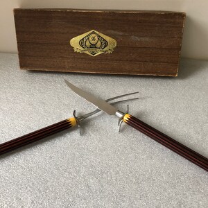 May include: A set of two carving tools with black and yellow handles. The tools are a knife and a fork. The tools are in a wooden box with a gold label that reads "The Connoisseurs Choice".