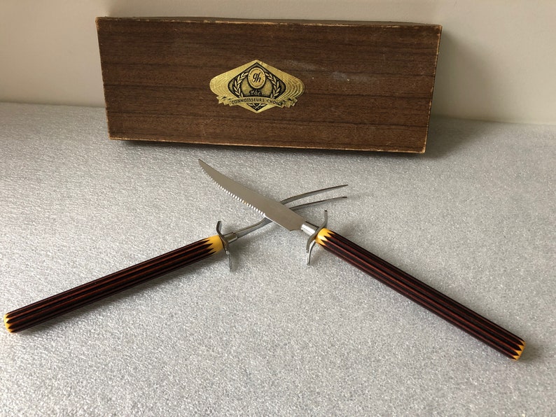 May include: A set of two carving tools with black and yellow handles. The tools are a knife and a fork. The tools are in a wooden box with a gold label that reads "The Connoisseurs Choice".