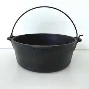 May include: A black cast iron cooking pot with a curved metal handle. The pot has a rounded shape with a wide opening and two small handles on the sides. The pot is likely used for cooking over a fire or on a stovetop.