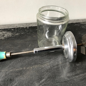 Peut inclure: Un bocal en verre vintage avec un couvercle en métal et un fouet en chrome. Le fouet a une poignée turquoise et blanche.