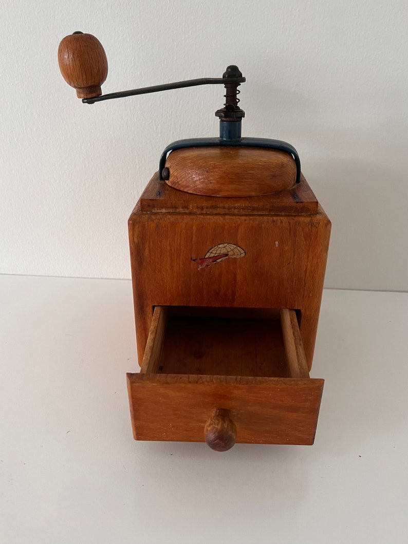 Puede incluir: Un molinillo de caf&eacute; de madera con cuerpo cuadrado, manivela de metal con pomo de madera y un caj&oacute;n extra&iacute;ble en la parte frontal. La parte superior tiene una tapa de madera y el frente muestra un peque&ntilde;o gr&aacute;fico de un globo terr&aacute;queo con avi&oacute;n.