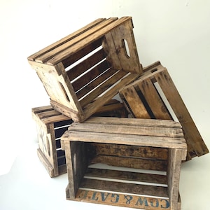 May include: A stack of four rectangular wooden crates with slatted sides and integrated hand-hold cutouts. The natural brown wood shows visible grain and weathering. One crate features the text COW & GATE printed in blue on the bottom slat.