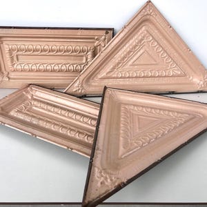 May include: Four vintage ceiling tiles with an ornate design. The tiles are made of metal and have a light pink color. Two of the tiles are triangular and two are rectangular.