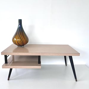 May include: A mid-century modern coffee table with a light beige top and a lower shelf. The table has black legs and a decorative glass vase with blue and amber swirls. The table is approximately 48 inches long.