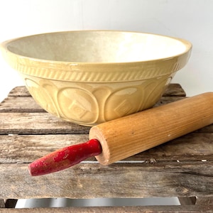 May include: A vintage-style beige ceramic mixing bowl with embossed geometric patterns and a wooden rolling pin with red handles. The bowl and rolling pin are displayed on a rustic wooden crate, creating a charming kitchen scene.