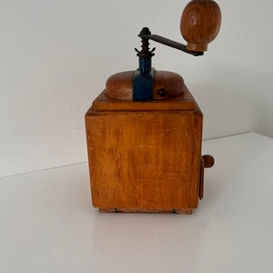 Puede incluir: Un molinillo de caf&eacute; cuadrado de madera con acabado marr&oacute;n claro, una placa superior de metal azul oscuro y una manivela manual con un pomo redondo de madera. El dispositivo incluye un peque&ntilde;o caj&oacute;n de madera con un pomo en la parte frontal.