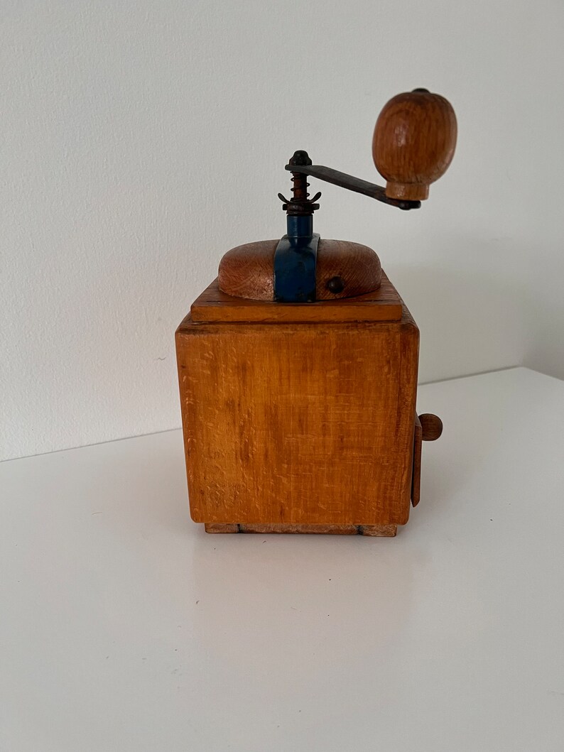 Puede incluir: Un molinillo de caf&eacute; cuadrado de madera con acabado marr&oacute;n claro, una placa superior de metal azul oscuro y una manivela manual con un pomo redondo de madera. El dispositivo incluye un peque&ntilde;o caj&oacute;n de madera con un pomo en la parte frontal.