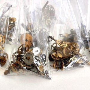 May include: Several clear plastic bags filled with various sizes of antique clock gears. The gears are made of brass and other metals, with a range of shapes and sizes. The bags are arranged closely together.