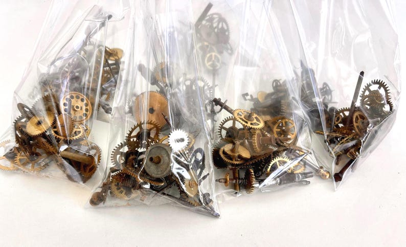 May include: Several clear plastic bags filled with various sizes of antique clock gears. The gears are made of brass and other metals, with a range of shapes and sizes. The bags are arranged closely together.