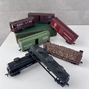May include: A collection of vintage toy train cars, including a boxcar labelled "Soo Line", a boxcar labelled "British Columbia Railway", a boxcar labelled "Delaware & Hudson", a flatbed car with logs, and two black tank cars.