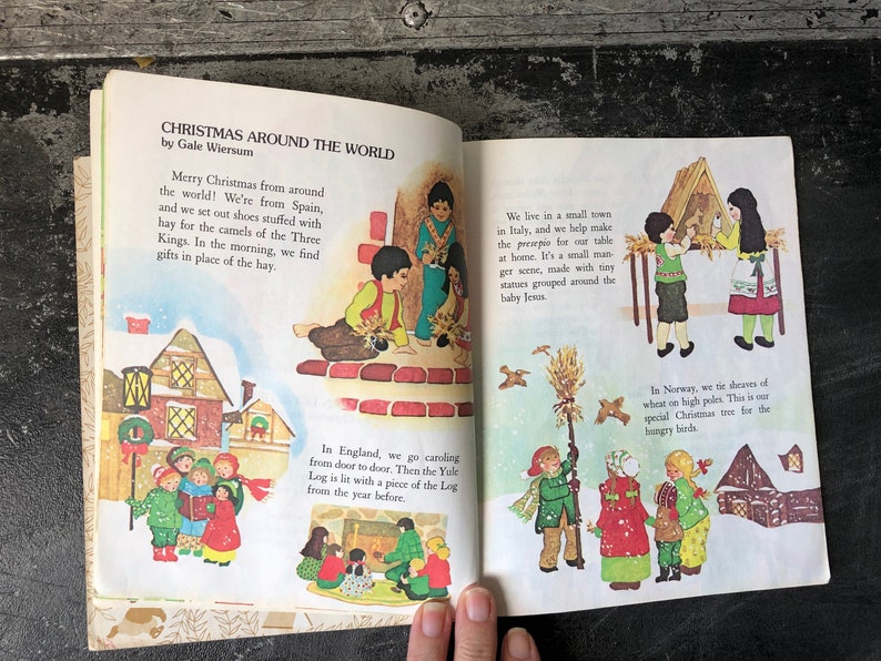 May include: An illustrated children's book page showing Christmas traditions around the world. The page features illustrations of children in Spain, Italy, England, and Norway, each celebrating Christmas in their own unique way. The text describes the traditions, such as leaving hay for the camels of the Three Kings in Spain, making a manger scene in Italy, caroling in England, and putting up sheaves of wheat for birds in Norway.