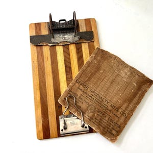 May include: A vintage wooden clipboard with a metal clip and a sheet of paper. The clipboard has a striped pattern in shades of brown. The paper has text that reads "OFFICE SPECIALTY MFG. CO."